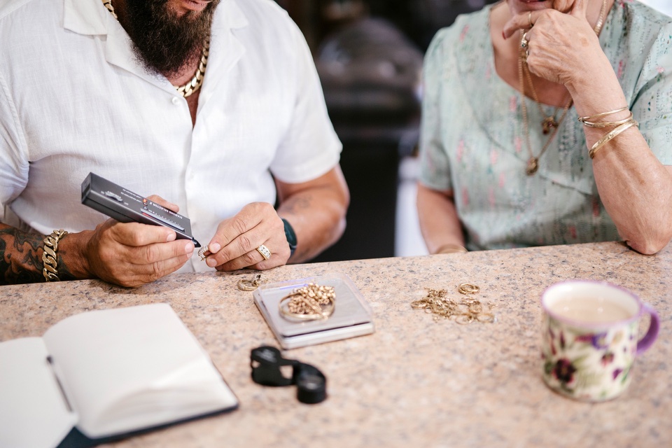 Jewellery valuation in-home session with a man using a diamond selector, an elderly woman observing, gold items on the table, and a cup of tea.