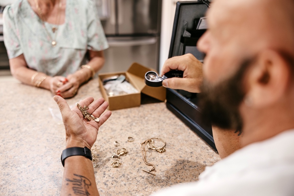 Man examining gold jewelry with loupe, woman in background, box of items on table, context of gold valuation and buying process.